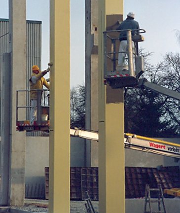 Maler stehen auf einer Baustelle auf Hubarbeitsbühnen und bringen Beschichtungen an Betonpfeilern an.